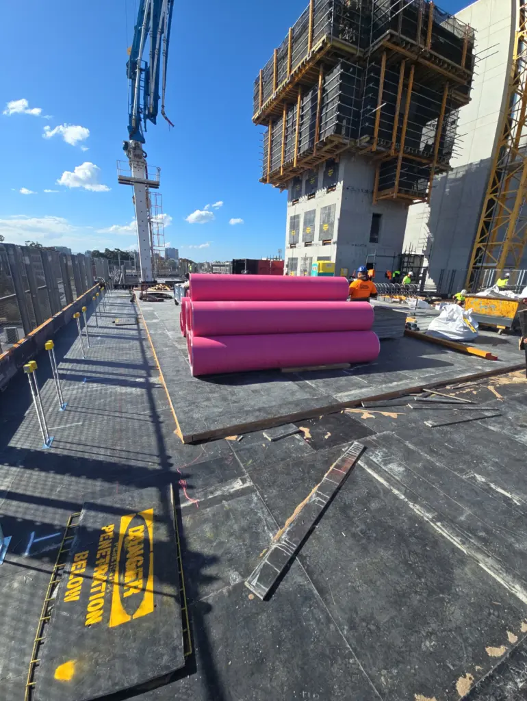 Stack of large bright pink pipes at a high-rise construction site with scaffolding and a concrete pump boom
