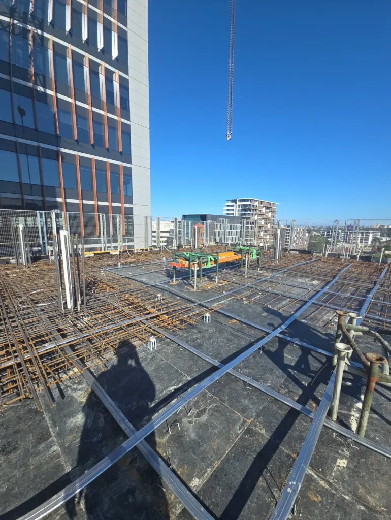 Rooftop construction site with rebar mesh and steel framing under a clear blue sky next to a high-rise building